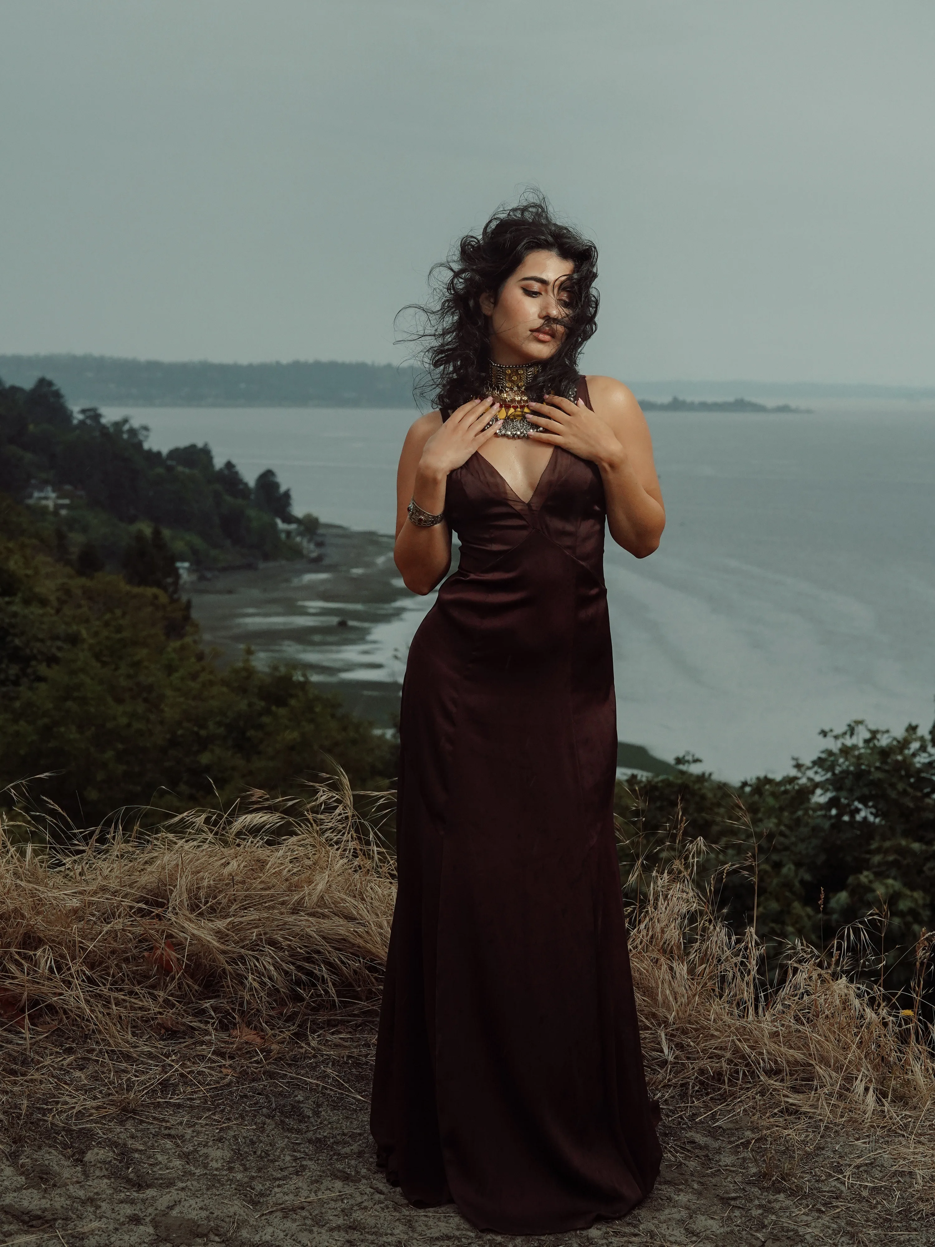 Sahra in a brown satin gown and statement necklace, hair in the wind, on a grassy bluff above the water